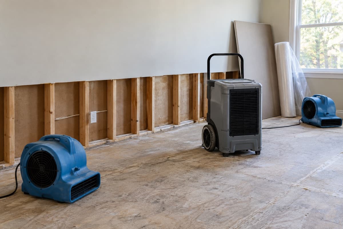 Air movers and a dehumidifier dry a room where the lower drywall has been removed, with new drywall and plastic sheeting ready for rebuilding
